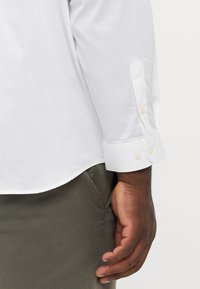 Chemise blanche à manches longues avec des poignets pointus ornés de deux boutons en perles, associée à un pantalon en tissu vert olive. Texture lisse et coupe ajustée.