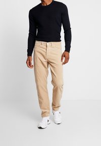 Beżowe spodnie sztruksowe o prostym kroju, zestawione z granatową prążkowaną koszulką z długim rękawem i białymi trampkami.