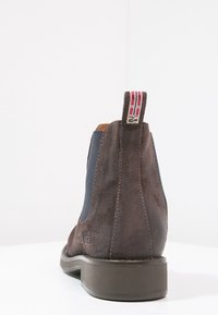 Bruine suède Chelsea-boot met marineblauwe zijpanelen, een lus met rood en witte strepen, en een lage rubberen hak. Textuurafwerking en stikseldetails.