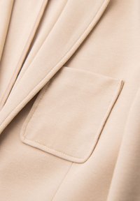 Gros plan sur un tissu beige avec une poche plaquée rectangulaire cousue et des coutures visibles le long de la poche et des bords du vêtement.