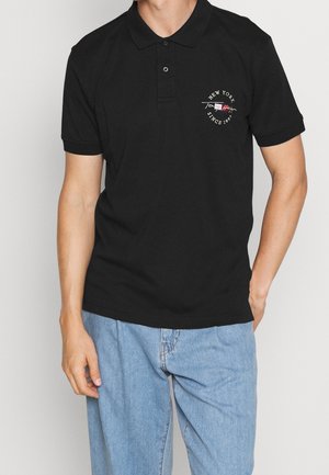 Czarna koszulka polo z klasycznym kołnierzykiem, krótkimi rękawami i małym wyhaftowanym logo na lewej piersi. Zestawiona z jasnoniebieskimi dżinsami.