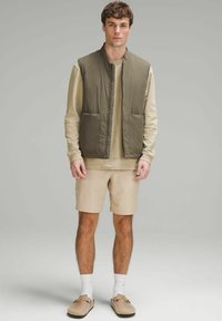 Khaki krāsa bez piedurknēm veste ar divām priekšējām kabatām, valkāta virs bēša garajām piedurknēm krekla, kopā ar bēša šortiem un gaišas krāsas apaviem.