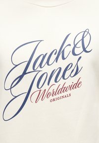 Biela tkanina s modrým nápisom "Jack & Jones" a červeným kurzívou napísaným "Worldwide Originals".