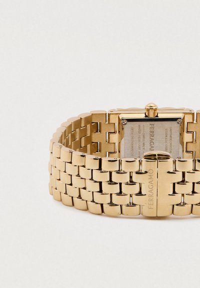 Zlaté kovové náramkové hodinky s obdĺžnikovým ciferníkom, spojeným remienkom a zapínaním na sponu. Na remienku je vygravírovaná značka a na zadnej strane hodiniek sú viditeľné detaily.