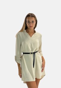 Lichtbeige, jurk met lange mouwen en een V-hals, getailleerd met een zwarte riem. Zachte textuur en een casual silhouet met opgerolde mouwen.