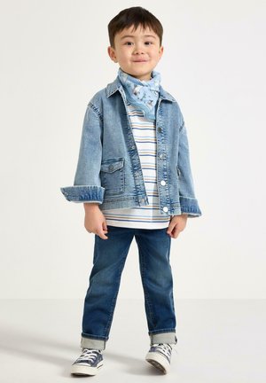 Młody chłopiec stoi i uśmiecha się, ubrany w jasną kurtkę dżinsową, pasiastą koszulę, niebieskie dżinsy, niebieskie trampki oraz jasnoniebieską wzorzystą chustę na szyi.