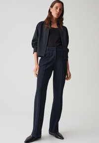 Donkerblauwe op maat gemaakte broek met voorplooien en zijsstripes, gecombineerd met een zwarte top en een zwarte ribbeltrui. Gladde stof textuur.