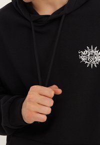 Černá mikina s kapucí s texturovaným povrchem, na hrudi s bílým grafickým designem "Mystic Whisper" a šňůrkami na kapuci.