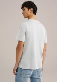 Balta medvilnė marškinėliai trumpomis rankovėmis, apvaliu iškirpimu ir nebaigtu kraštu. Dėvimi su šviesiai mėlynomis džinsinėmis kelnėmis, demonstruojančiomis kasdienį stilių.