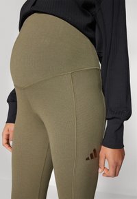 Ελιά πράσινες ραβδωτές κολάν με ψηλή μέση. Διαθέτει ένα σκούρο καφέ λογότυπο Adidas στο πλάι και υφασμάτινη υφή, σε συνδυασμό με ένα μαύρο τοπ.