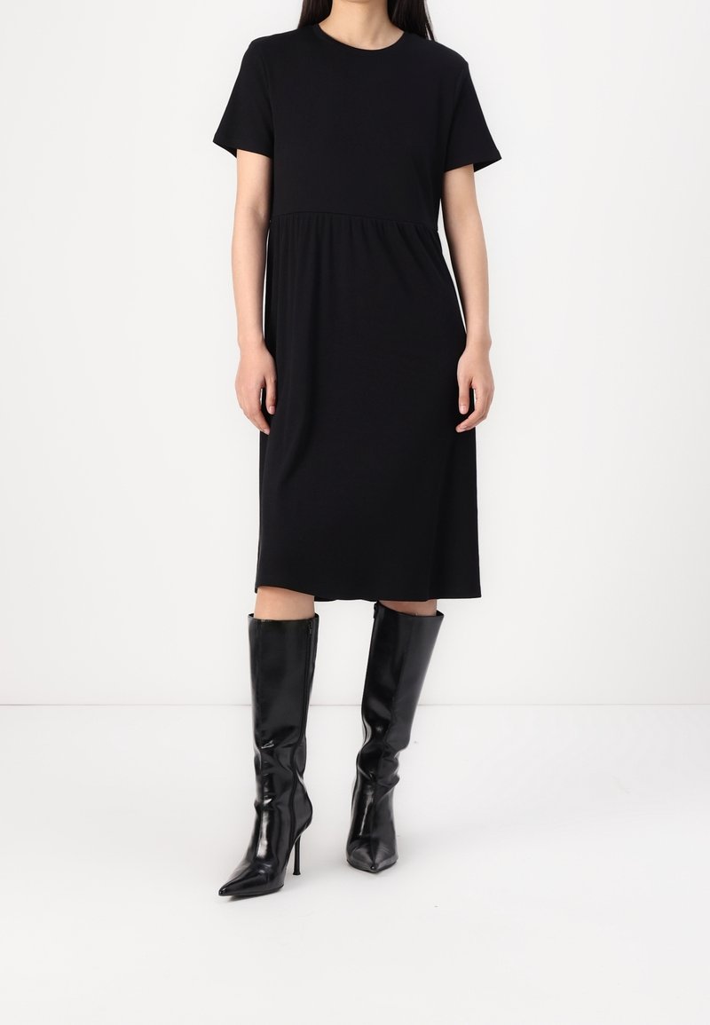 Robe t-shirt noire à manches courtes, coupe décontractée et longueur mi-mollet. Portée avec des bottes noires hautes à talons aiguilles au fini brillant.