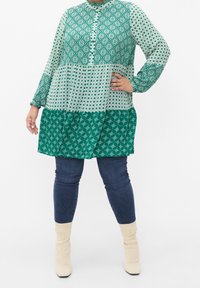 Tunika verde a fantasia con maniche lunghe, caratterizzata da design geometrici in diverse tonalità di verde. Taglio A-line con chiusura a bottoni. Jeans in denim e stivali beige.
