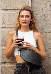 Junge Frau mit langen, lockigen Haaren, die ein weißes Crop-Top und schwarze Jeans trägt, hält einen Becher mit Eiskaffee vor einem Steinwand-Hintergrund.