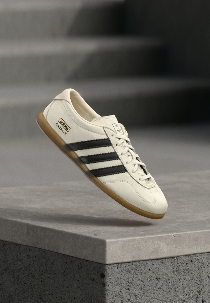 Λευκό δερμάτινο αθλητικό παπούτσι με τρεις μαύρες ρίγες, στρογγυλή μύτη και σόλα από καουτσούκ. Λογότυπο Adidas στο πλευρό και σήμανση «Gazelle» στη φτέρνα.