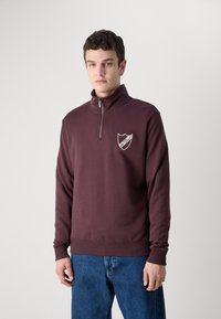 Bordo flis pulover s polu zatvaračem, rebrastim manžetama i rubom. Ima mali grb logotip na lijevoj strani prsima. Mekana, glatka tekstura.
