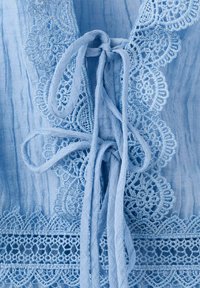 Tissu bleu clair avec délicate bordure en dentelle et nœuds en tissu noués au centre, montrant une texture et un motif détaillés.