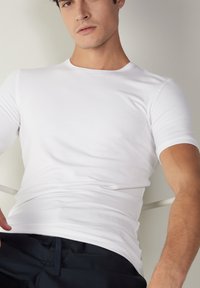 T-shirt blanc à manches courtes en tissu coton lisse, avec un col rond et une coupe ajustée, associé à un pantalon sombre.