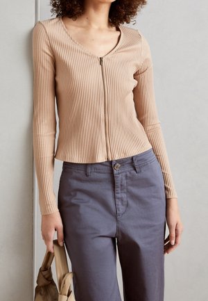 Beżowy prążkowany top z długim rękawem, dekoltem w serek i przednim zamkiem błyskawicznym, zestawiony z szarymi spodniami z zapięciem na guzik i bocznymi kieszeniami.