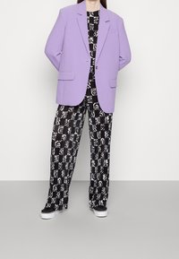 Lila oversize marynarka na czarnym i białym wzorzystym komplecie z szerokimi spodniami. Sneakersy dopełniają look, z białymi podeszwami i czarnymi akcentami.