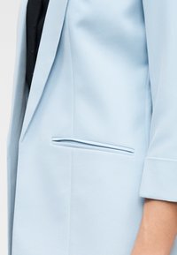 Μανίκι ανοιχτό μπλε σακακιού και λεπτομέρεια τσέπης πάνω από μαύρο ρούχο στο μπράτσο και τον κορμό ενός ατόμου.