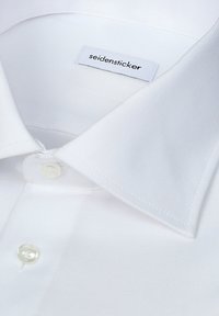 Col de chemise blanche et boutons supérieurs avec étiquette de marque "seidensticker" visible sur tissu texturé.