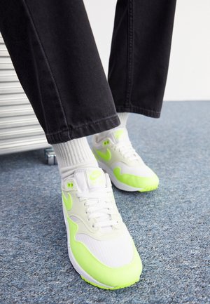 Person iført hvide og neongrønne Nike Air Max sneakers, hvide sokker og sorte bukser står på blåt tæppe.