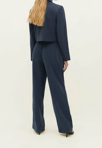 Laivastonsininen räätälöity puku, jossa on lyhyt takki ja leveälahkeiset housut. Sileä, teksturoitu kangas, minimaalinen yksityiskoristus ja tyylikäs istuvuus.