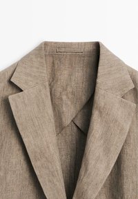 Close-up van de kraag en revers van een lichtbruine linnen blazer, waarbij de stoftextuur en stikdetails zichtbaar zijn.