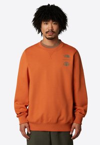 Orange sweatshirt i mjukt tyg, har rundad hals, ribbade muddärmar och två logotyper i tealtryck på bröstet för kontrast.