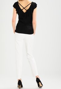 Žena v černém topu s kříženými ramínky a volánkovým zadním dílem, bílých kalhotách a černých vysokých podpatcích, stojící s rukama v kapsách.