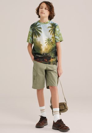 Băiat purtând un tricou cu imprimeu tropical cu palmieri și o jeep, pantaloni scurți cargo kaki, șosete albe, adidași maro și ținând o geantă mică verde.
