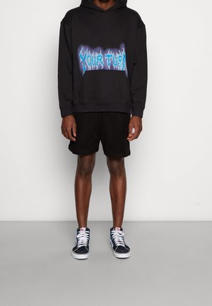 Černá mikina s kapucí a grafickým textem, v kombinaci s černými síťovanými šortkami. Obuté s modro-bílými teniskami, vytvářející ležérní streetwear styl.