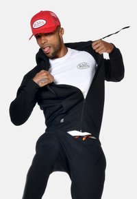 Uomo con cappello rosso, felpa nera con zip sopra una maglietta bianca con logo Von Dutch, che apre la felpa e tira fuori la lingua, su uno sfondo uniforme.