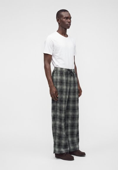 Homme debout portant un t-shirt blanc uni à manches courtes, un pantalon large à carreaux gris et noir, et des chaussures marron foncé sur fond blanc.