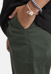 Een hand van een persoon in de voorzak van een donkergroen broek, draagend een zwart shirt en twee armbanden, één roze met een witte sluiting en één met een blauw patroon.