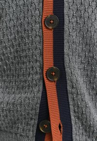 Grå texturerad tröja med ett stickat mönster, orange och marinrand ner längs sidan, och mörka knappar för stängning.