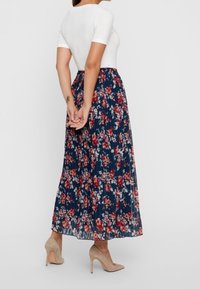 Kvinna klädd i vit kortärmad topp, marinblå blommig plisserad maxikjol och beige högklackade skor, står med händerna ihopklämda bakom ryggen.