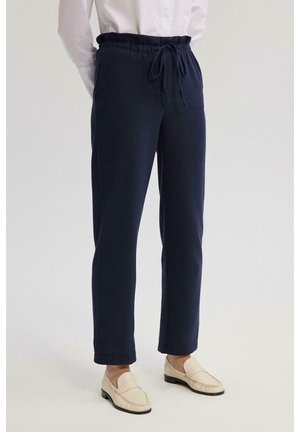 Navy blauwe broek met elastische taille en trekkoord, taps toelopende snit en zachte textuur. Gecombineerd met crèmekleurige instappers op een lichte achtergrond.