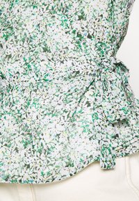 Blusa floreale in tessuto leggero con un motivo verde e bianco caratterizzato da piccoli fiori. Include un dettaglio con nodo laterale in vita.