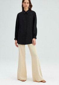 Zwarte oversized shirt met een kraag, lange mouwen en knoopsluiting aan de voorkant, gecombineerd met brede beige broek en sandalen. Glad textuur.