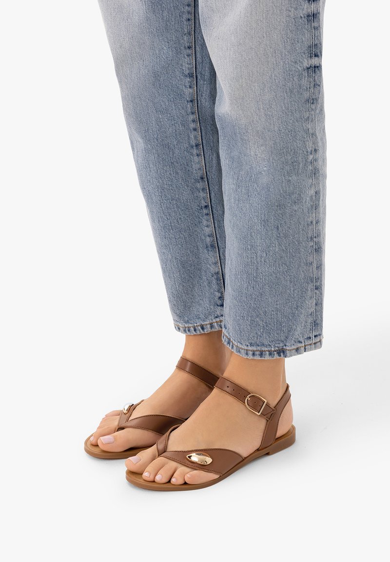 Pieds portant des sandales marron à bride à la cheville avec des accents dorés, associés à un jean denim bleu clair coupé au-dessus des chevilles.