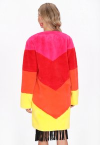 Manteau en fausse fourrure colorée présentant un motif en chevrons rose, rouge, orange et jaune, avec des manches longues et un ourlet à franges.