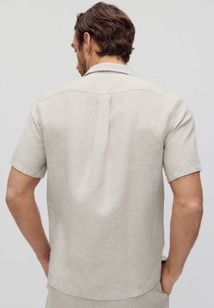 Mężczyzna o brązowych falowanych włosach, ubrany w jasnobeżową lnianą koszulę z krótkim rękawem, stoi tyłem, z rękami przy kieszeniach, na jednolitym tle.