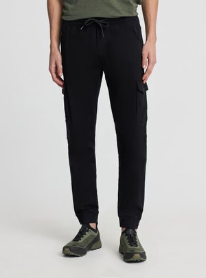 Persona che indossa pantaloni jogger cargo neri con vita con coulisse e scarpe da trekking verde oliva e nere, in piedi su sfondo uniforme.