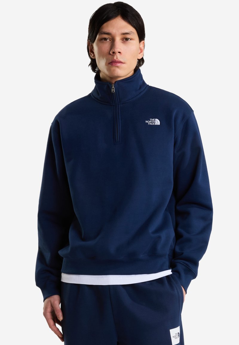 Joven con cabello oscuro que lleva un pulóver North Face azul marino con cremallera hasta el pecho y pantalones a juego, posando delante de un fondo liso.