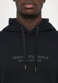 Hombre con sudadera negra con capucha que presenta el texto "ARMANI EXCHANGE MILANO / NEW YORK" y cuerdas negras con extremos de metal visibles.