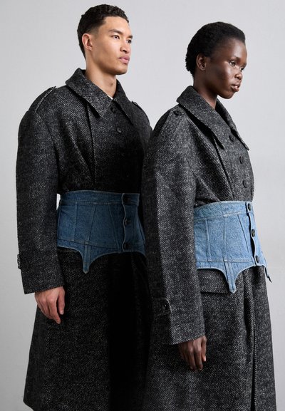 Dos modelos llevan abrigos largos de espiga oscuros con acentos de corsé de mezclilla azul en contraste y grandes cuellos, con detalles de botones.