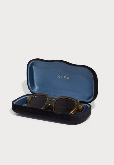 Gucci napszemüveg fekete bársonytokban, kék belsővel. A szemüveg átlátszó arany kerettel és sötét lencsékkel rendelkezik. Arany logó látható.
