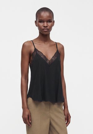 Donna con capelli corti che indossa un top nero con rifiniture in pizzo e pantaloni beige, in piedi davanti a uno sfondo bianco semplice.