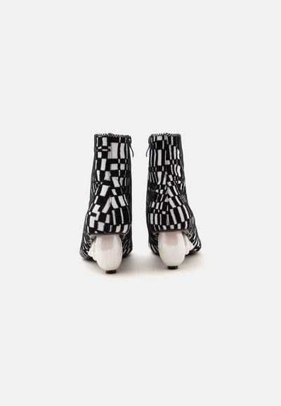 Bottines à motifs noir et blanc avec talons blancs incurvés uniques, vues de dos sur un fond clair uni.